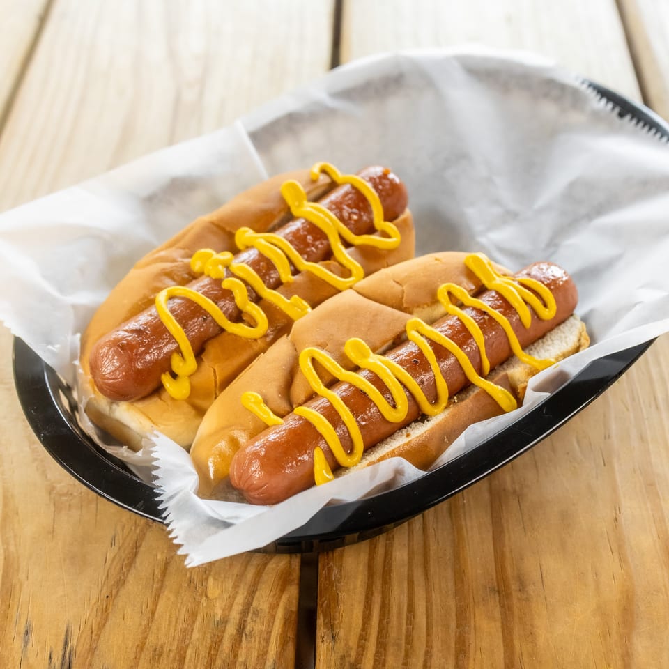 Two hot dogs in toasted buns topped with yellow mustard, served in a paper-lined basket on a wooden table.