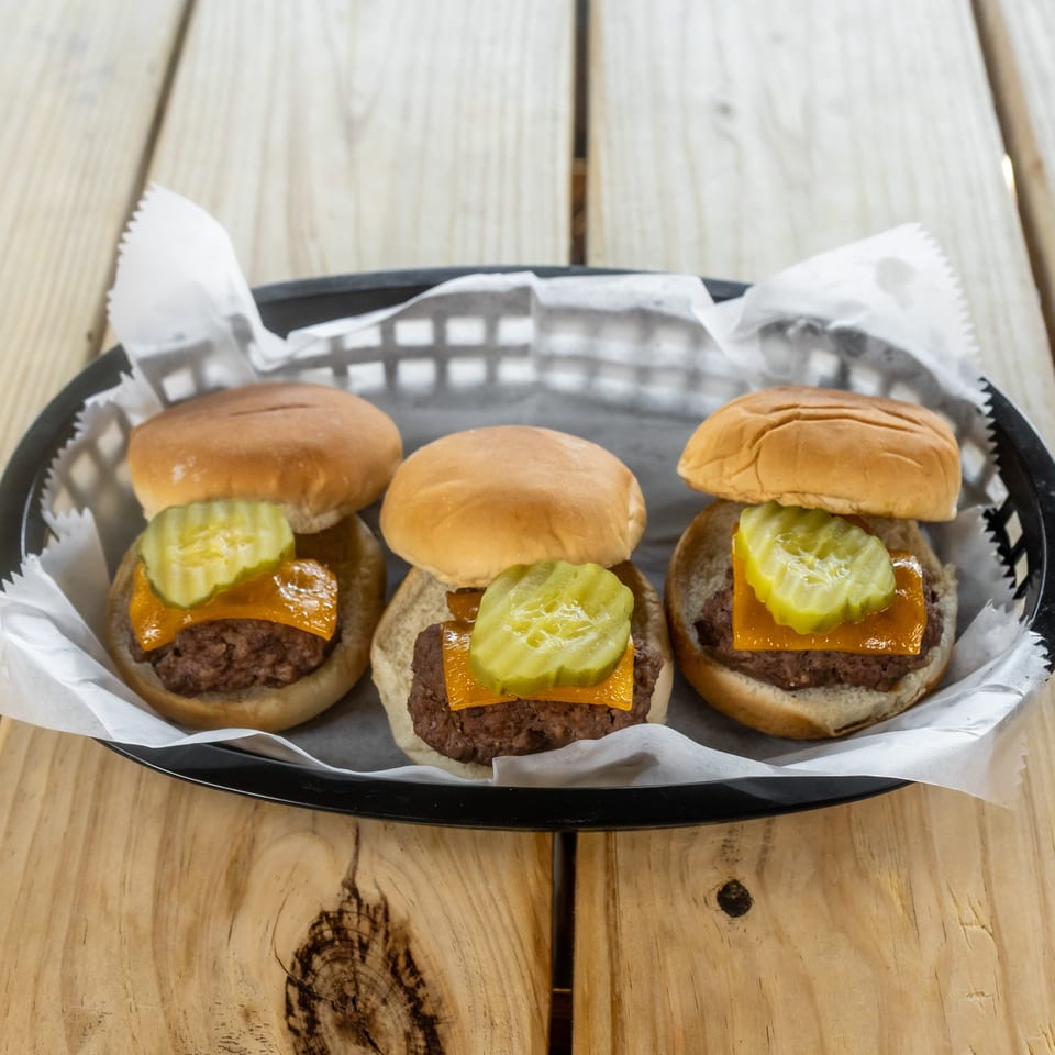 Three cheeseburgers topped with sliced pickles on a paper-lined black tray atop a wooden table.