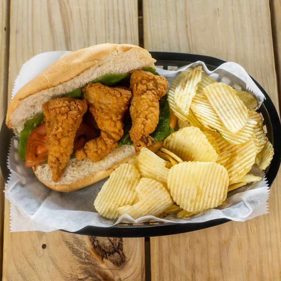 Fried chicken sandwich with lettuce and tomato served alongside ridged potato chips in a paper-lined black basket.