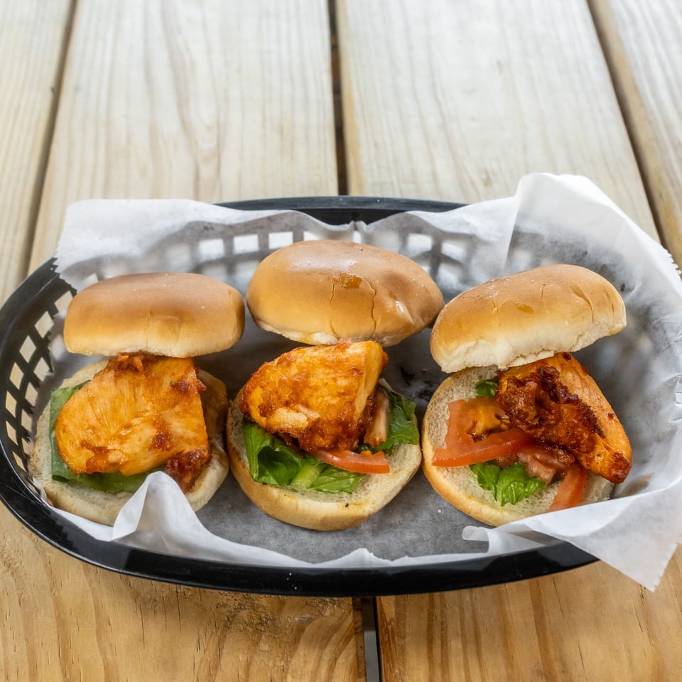 Three crispy chicken sandwiches with lettuce, tomato, and bacon served in a lined basket on a wooden table.