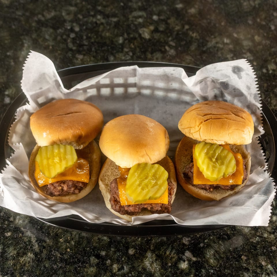 Three cheeseburgers topped with pickles on soft buns, arranged in a paper-lined black tray on a granite countertop.