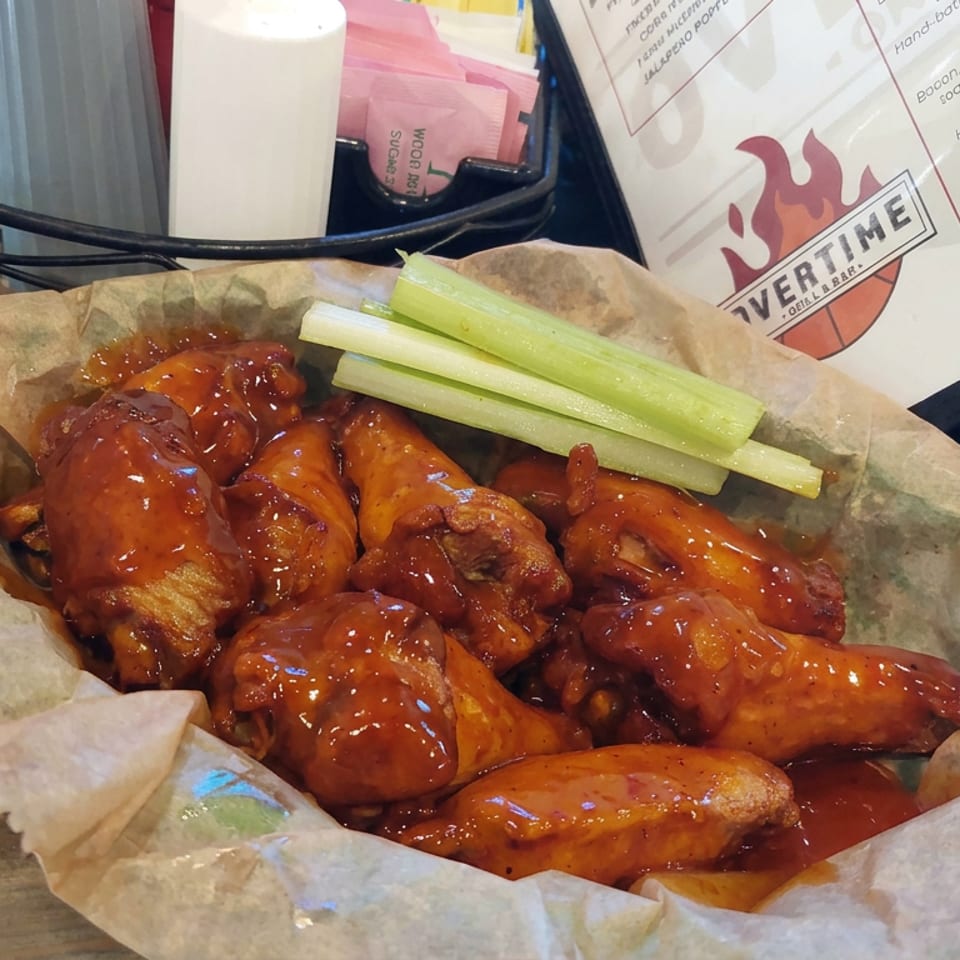 Basket of glossy red-sauced chicken wings with celery sticks on parchment paper, beside a menu and table condiments.