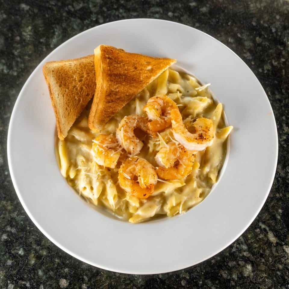 Creamy penne pasta topped with seasoned shrimp, grated cheese, and toasted bread triangles on a white plate.