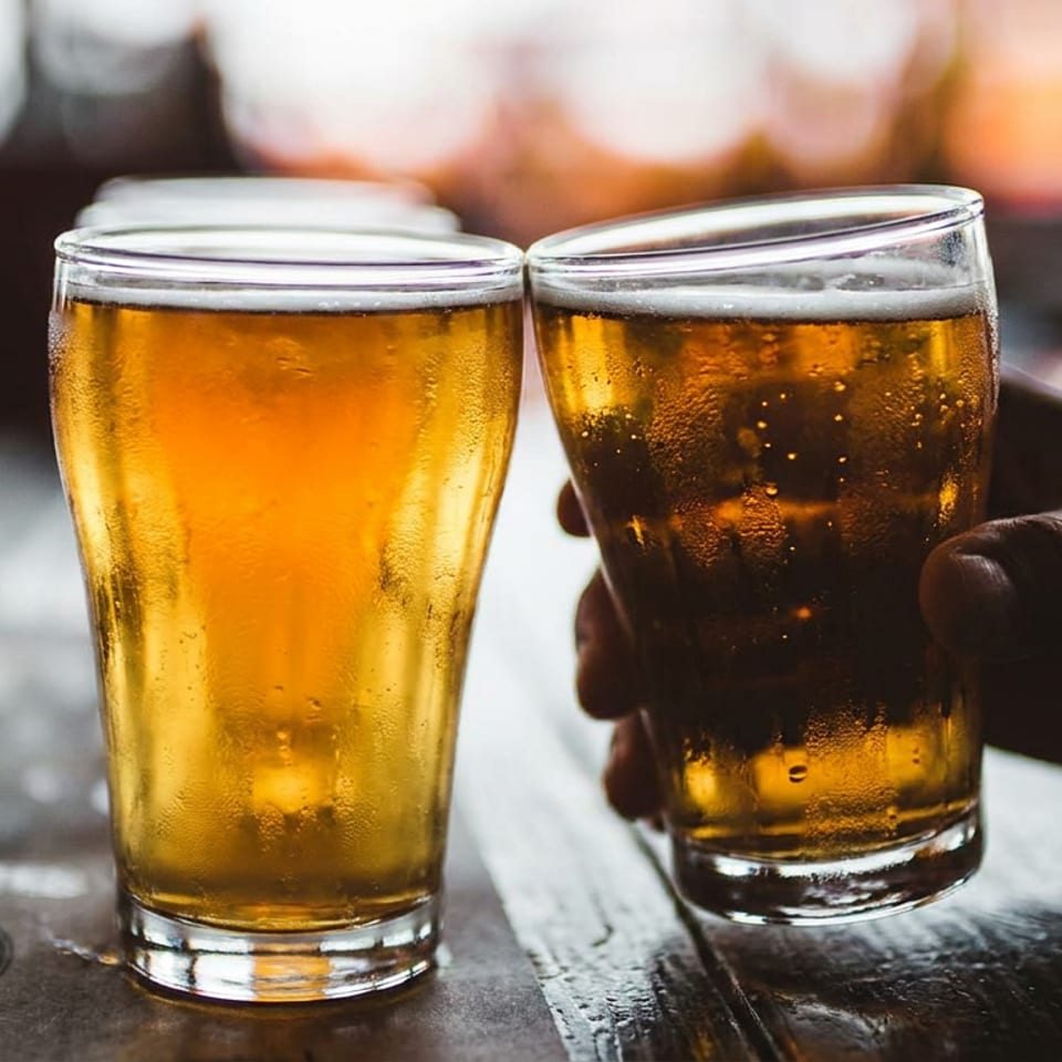 Two frosty beer glasses sit on a rustic wooden table, one held by a hand, with a warmly lit bar blurred in the background.