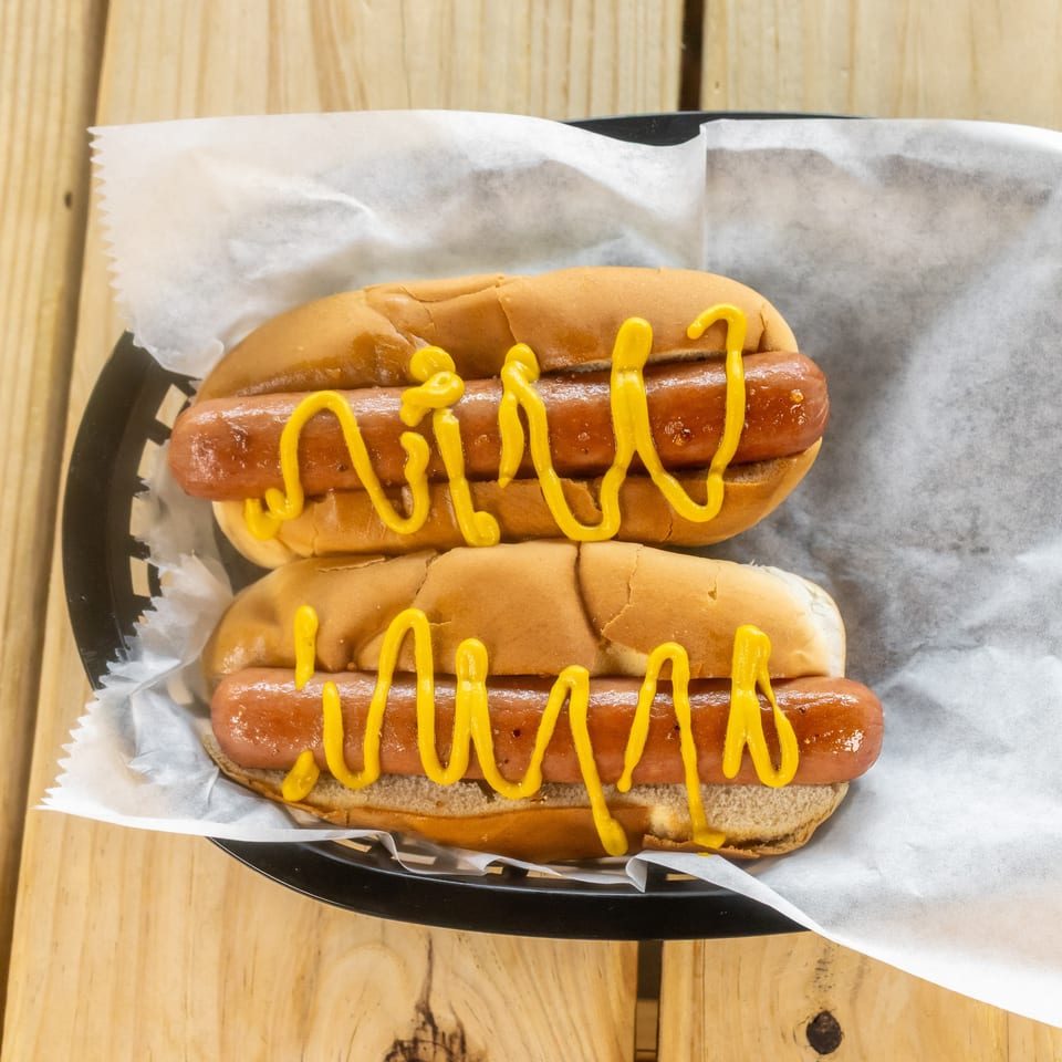 Two grilled hot dogs in buns topped with yellow mustard, served on wax paper in a black tray.