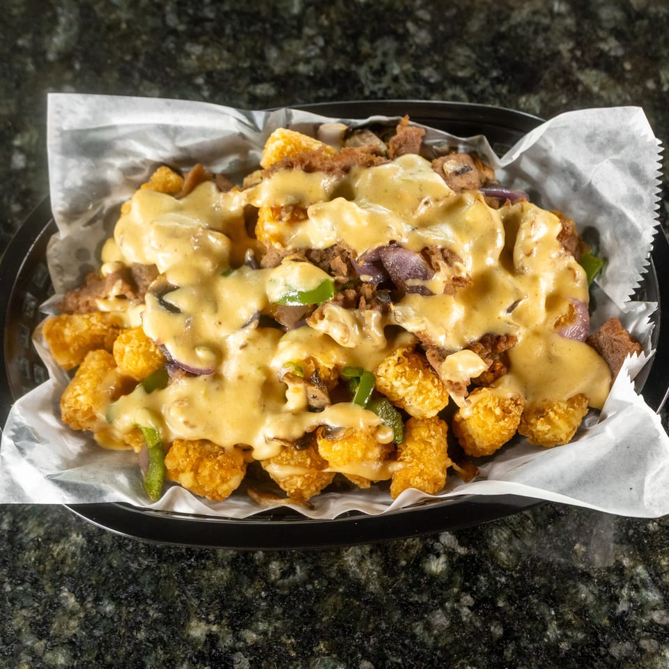 Crispy tater tots topped with melted cheese sauce, fried onions, mushrooms, and green peppers in a lined serving tray.
